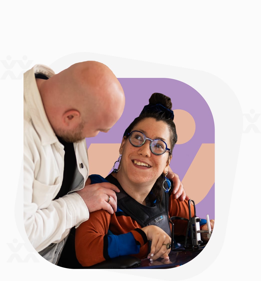 Een man buigt zich lachend over een vrouw in een rolstoel die aan een tafel werkt.