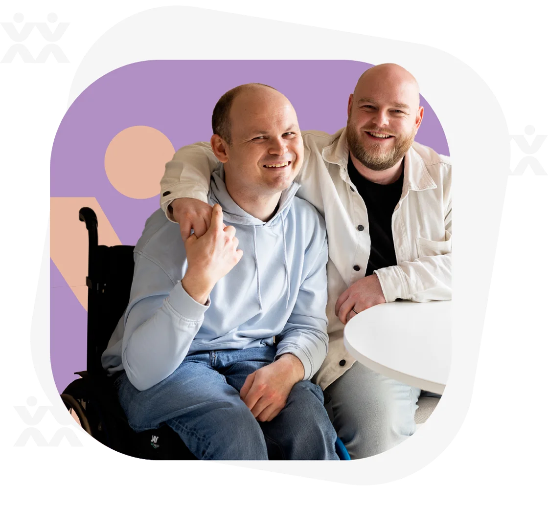 Twee vrienden, waarvan één in een rolstoel, lachen samen aan een tafel.